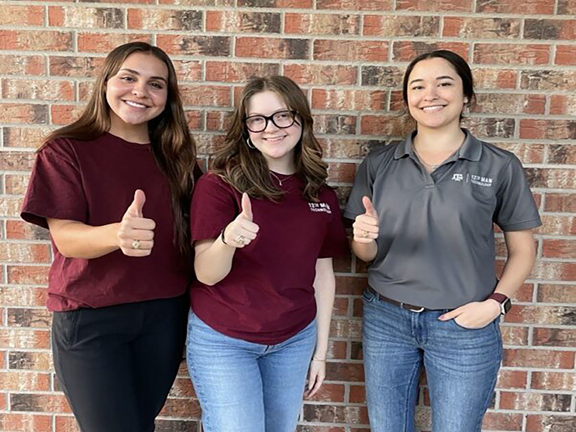 3 students in front of a brick wall with thumbs up.